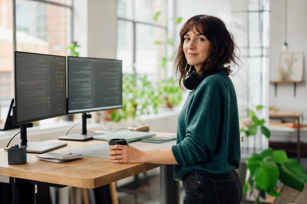 A developer at a standing desk with two screens, one hand on a coffee cup, plants in the window
