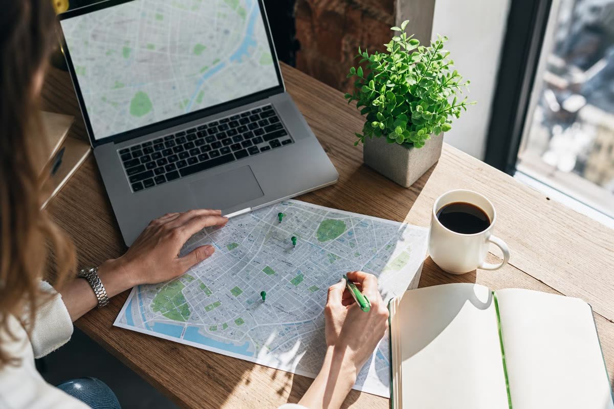 A desk by a window with a laptop, a printed city map with green pins, and a hand marking a spot