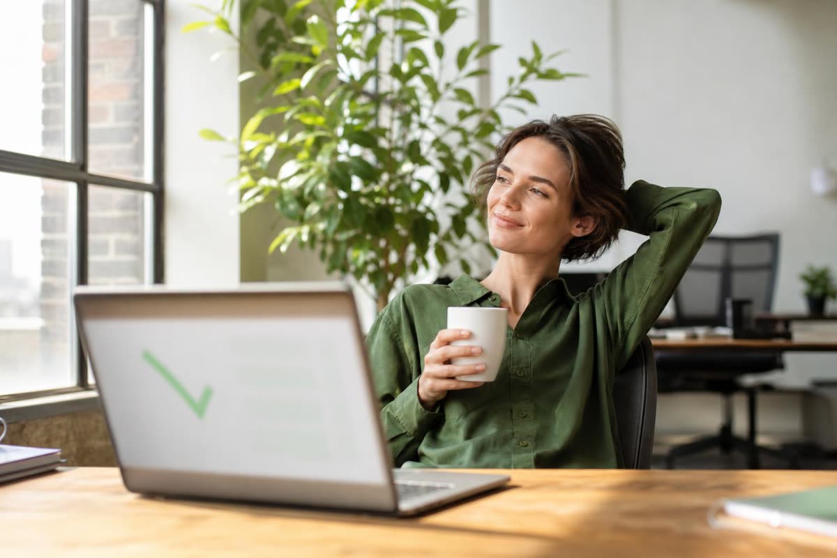A consultant leaning back with a coffee, a green tick on the laptop in front of her